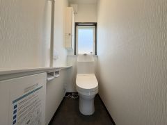 シンプルで使いやすいトイレ空間です。窓が設けられているため換気がしやすい設計となっています。白を基調とした内装が清潔感を演出しています。手すりや小物置きスペースがあり機能性にも配慮されています。