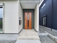 玄関はシンプルながらも洗練されたデザインです。木目調のドアが落ち着いた雰囲気を演出します。タイル敷きのポーチが玄関まわりを印象的に仕上げています。外壁とのコントラストも際立ちます。