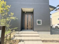 重厚感のある玄関扉が建物全体の雰囲気に調和しています。ステップやアプローチ部分も広々としており開放感を感じられます。外壁のシックなカラーと木目の扉がモダンな印象を与えます。